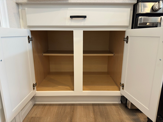 An empty white kitchen cabinet with two open doors and a single shelf inside, set against a wooden floor. The cabinet is under a countertop and next to a stainless steel oven.