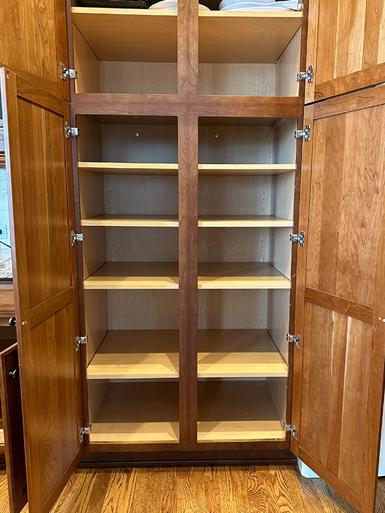 A large wooden pantry cabinet with both doors open, revealing six empty shelves inside. The cabinet is made of light and medium brown wood and stands on a wood floor.