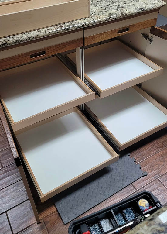 Four pull-out shelves are extended from a lower kitchen cabinet beneath a granite countertop. A black mat and an open organizer with tools and hardware are on the wooden floor nearby.