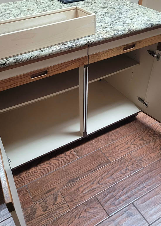 Open kitchen cabinet with two doors and empty shelves beneath a granite countertop; a wooden drawer box sits on top. The floor is dark wood with a visible grain pattern.