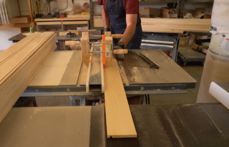 A person uses a table saw to cut a plank of wood in a workshop, wearing an apron and safety gear. Stacks of wood and various tools are visible in the background.