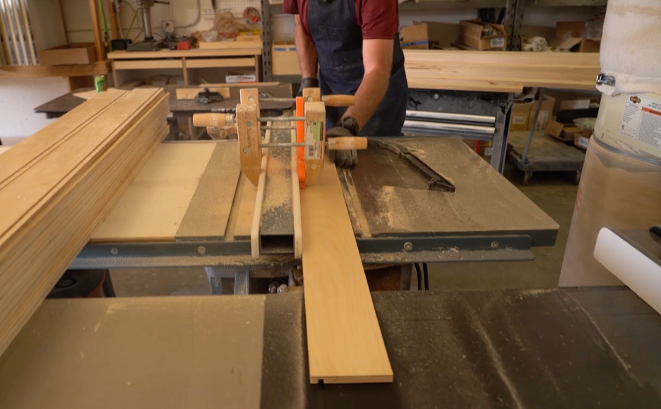 REOS workshop1 A person uses a table saw to cut a plank of wood in a workshop, wearing an apron and safety gear. Stacks of wood and various tools are visible in the background.