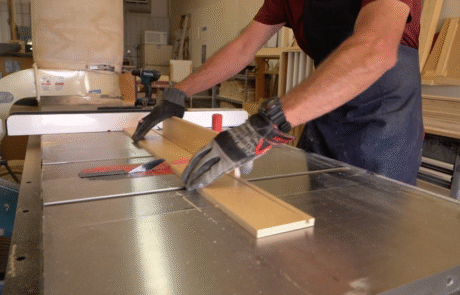 A person wearing gloves uses a table saw to cut a piece of wood in a workshop, with various tools and materials visible in the background.