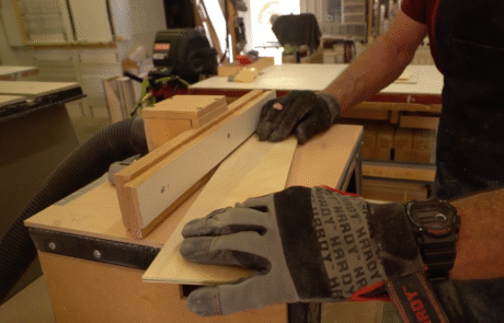 A person wearing work gloves guides a piece of wood through a table saw in a workshop, using a fence to ensure a straight cut. Tools and materials are visible in the background.