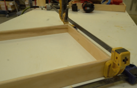 A wooden frame is being clamped together on a workbench, with glue visible at the joints. Tools and workshop items are seen in the background.