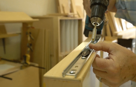A person uses a power drill to attach a metal drawer slide to the side of a wooden drawer in a woodworking shop.