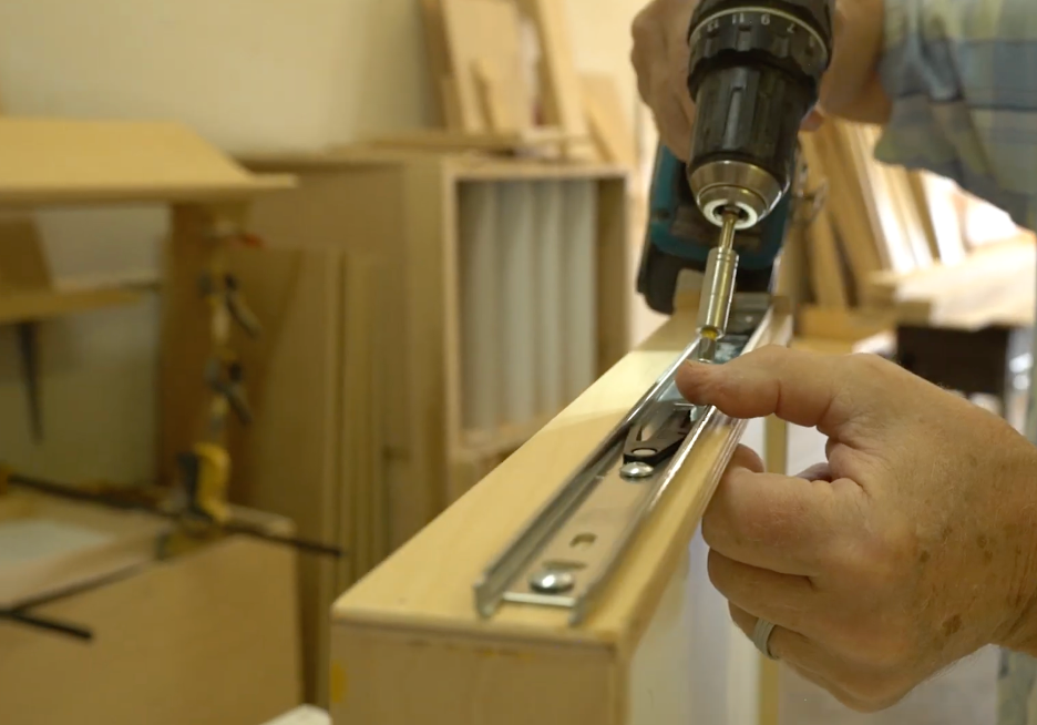 REOS workshop6 A person uses a power drill to attach a metal drawer slide to the side of a wooden drawer in a woodworking shop.