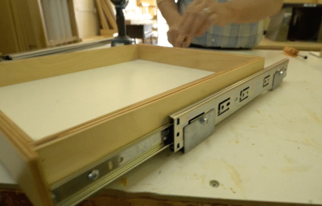 A person installs a drawer with metal slides and built-in electrical outlets on the side, in a woodworking shop. The drawer sits on a workbench among tools and wood pieces.