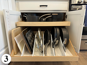 ba-3 A kitchen cabinet with two shelves: the top shelf holds pots and pans, while the bottom shelf has white dividers organizing baking sheets, cutting boards, muffin pans, and skillets. A number 3 is in the bottom left corner.