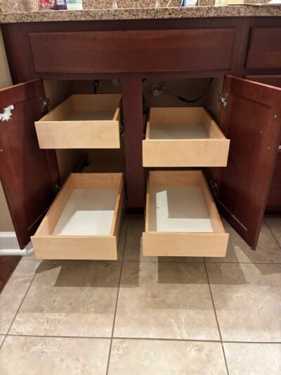 Four wooden drawers are pulled out from a bathroom vanity beneath a sink, with the cabinet doors open and plumbing visible. The drawers and cabinet are empty, and the floor is tiled.