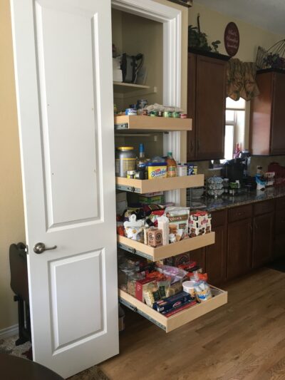 A white pantry door is open, revealing four pull-out shelves filled with various food items and groceries in a kitchen with wooden cabinets and a hardwood floor.