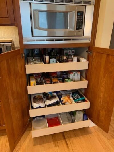 A kitchen cabinet with open wooden doors reveals several pull-out shelves stocked with pantry items, snacks, containers, and a microwave oven installed above the shelves.
