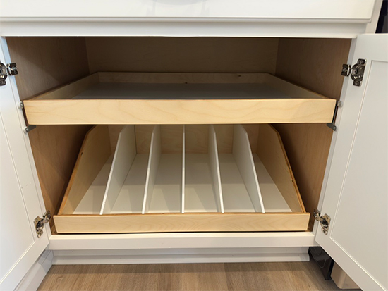 Open white cabinet showing two pull-out wooden shelves; the bottom shelf has vertical dividers for organizing items like baking sheets or trays, while the top shelf is a flat surface with raised edges.