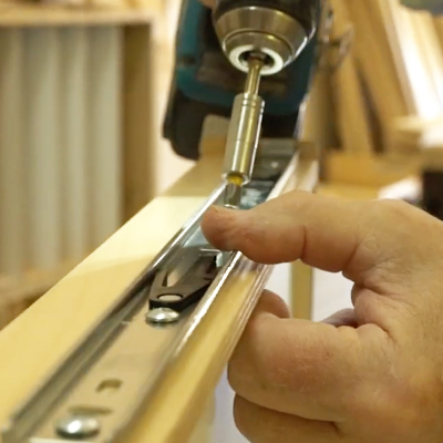 home-handcrafted-sized – Roll 'em Out Shelves A close-up of a person’s hand installing a metal drawer slide onto a wooden board using a power drill in a woodworking shop.