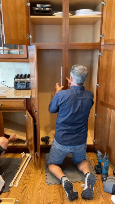 Installer kneeling on a mat, measuring the inside of a large wooden kitchen cabinet with a tape measure