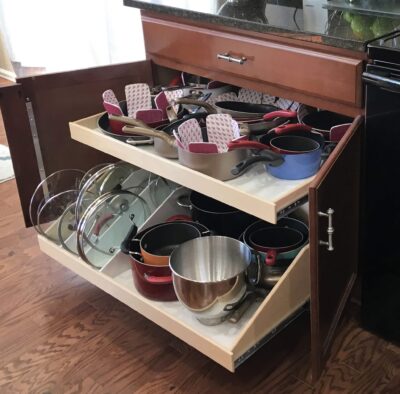 Open kitchen cabinet with two pull-out shelves neatly organized with pots, pans, lids, and baking trays. The shelves are wooden, and the cabinet is brown with metal handles. The kitchen floor is hardwood.