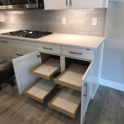 A modern kitchen with a gas stove, white cabinets, and two lower cabinet doors open to reveal pull-out wooden shelves on both sides. The floor is light-colored wood, and the walls have a light gray backsplash.