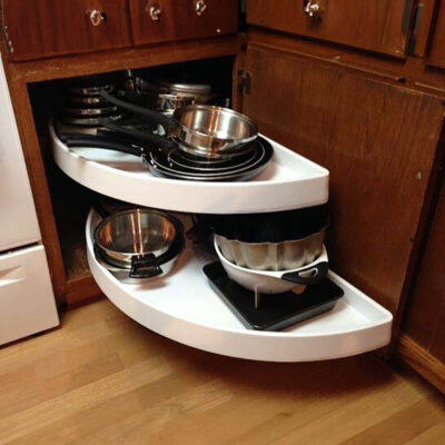 reos-product-gallery-26 – Roll 'em Out Shelves A corner kitchen cabinet with two white pull-out shelves, each holding various pots, pans, and bakeware, efficiently organized for easy access. The cabinet is made of dark wood and sits on a wooden floor.