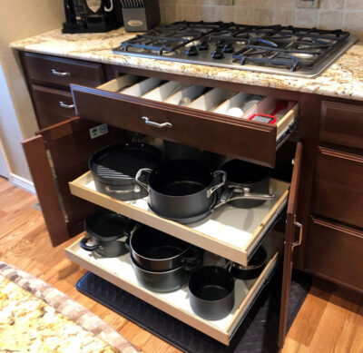 reos-product-gallery-30 – Roll 'em Out Shelves A kitchen cabinet with sliding drawers open, showing neatly organized pots, pans, and lids below a countertop with a gas stove and coffee maker.