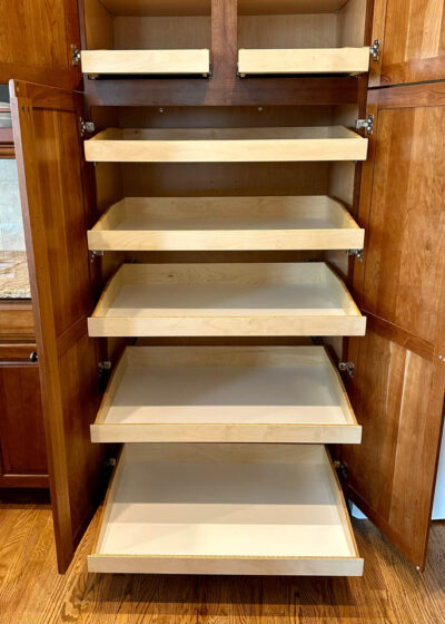 A wooden cabinet with double doors is open, revealing six pull-out shelves or drawers inside. The shelves are empty, and the cabinet is set on a hardwood floor.