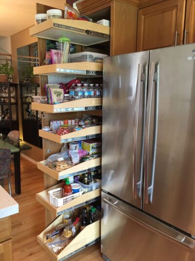 A tall wooden pull-out pantry with six shelves is fully extended next to a stainless steel refrigerator, showing various organized food items, condiments, bottled water, and snacks on each shelf.