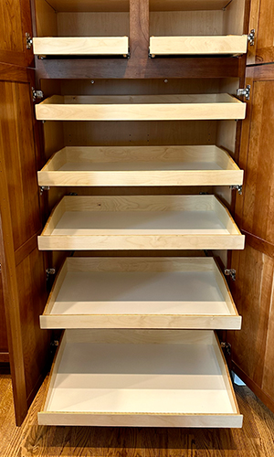 straightforward-new – Roll 'em Out Shelves A wooden pantry cabinet with its doors open, revealing six empty sliding shelves made of light-colored wood, all pulled out to different lengths. The cabinet sits on a hardwood floor.
