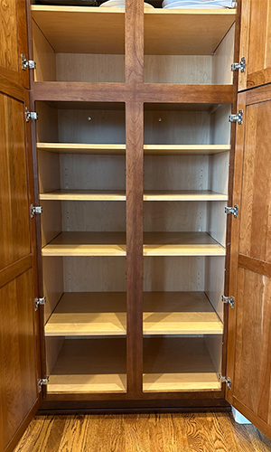straightforward-old – Roll 'em Out Shelves An empty wooden pantry cabinet with double doors is open, revealing six shelves inside. The wood has a warm brown finish, and the shelves and interior have a lighter color. The floor beneath is hardwood.